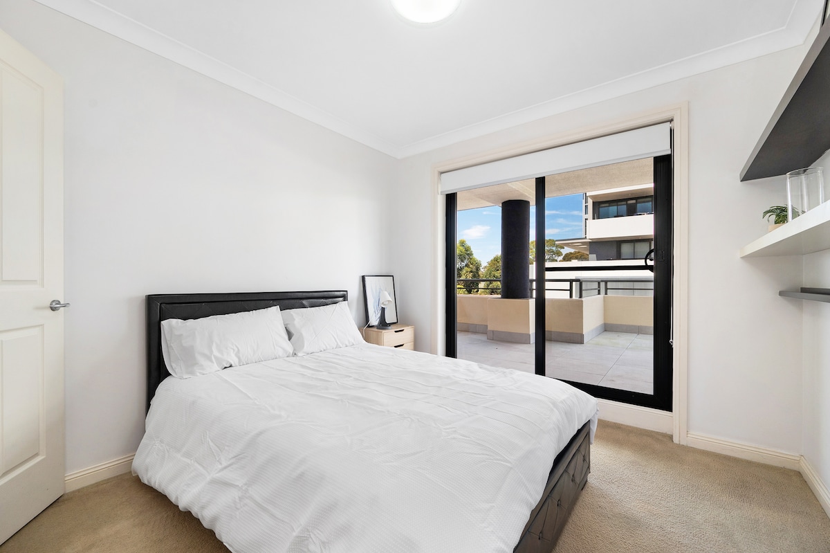 A well-lit bedroom features a spacious bed with neatly arranged bedding. Floor-to-ceiling glass doors provide a view of a balcony, while a minimalist shelf is installed on one wall. Light-colored walls contribute to a bright and airy ambiance.
