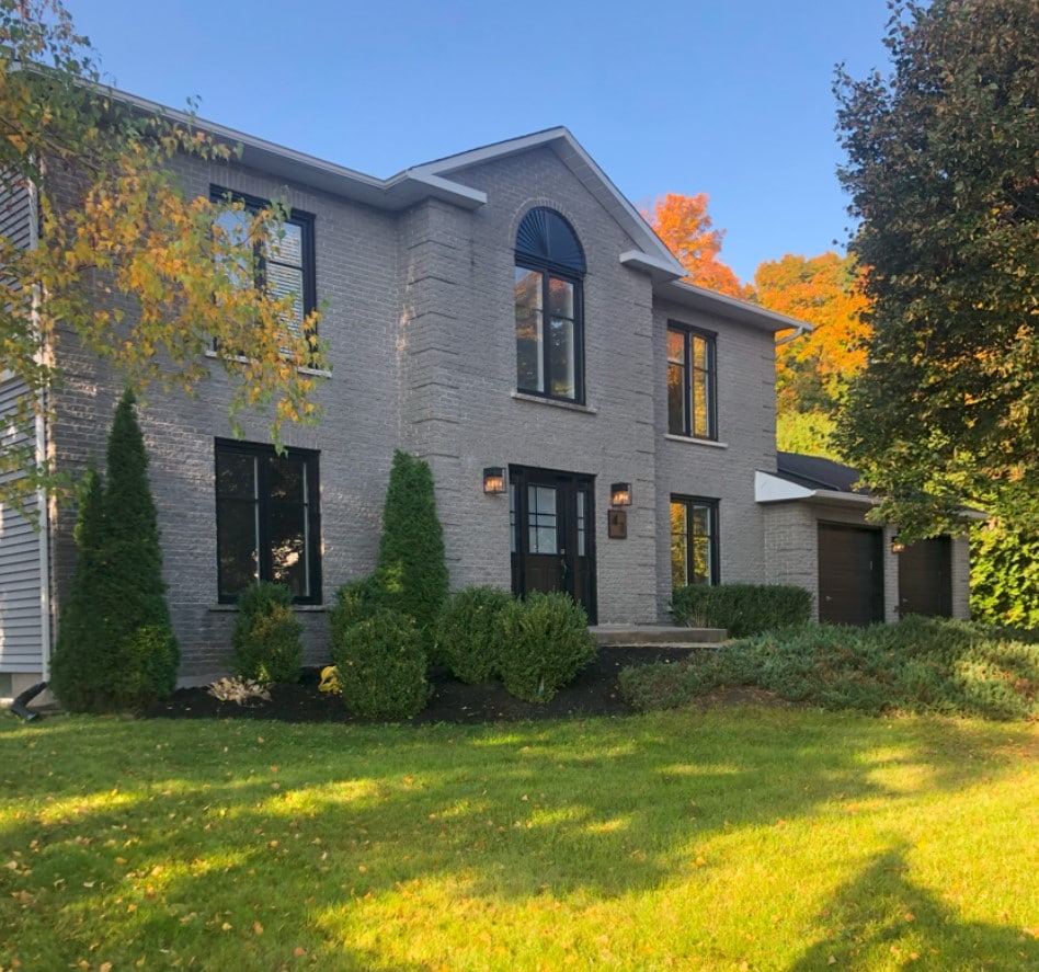A spacious two-story house with modern brick exterior and large windows is surrounded by manicured greenery. Flanked by neatly trimmed shrubs and trees, the entrance is characterized by a set of welcoming doors under an arched window, presenting a peaceful view of the residential area.