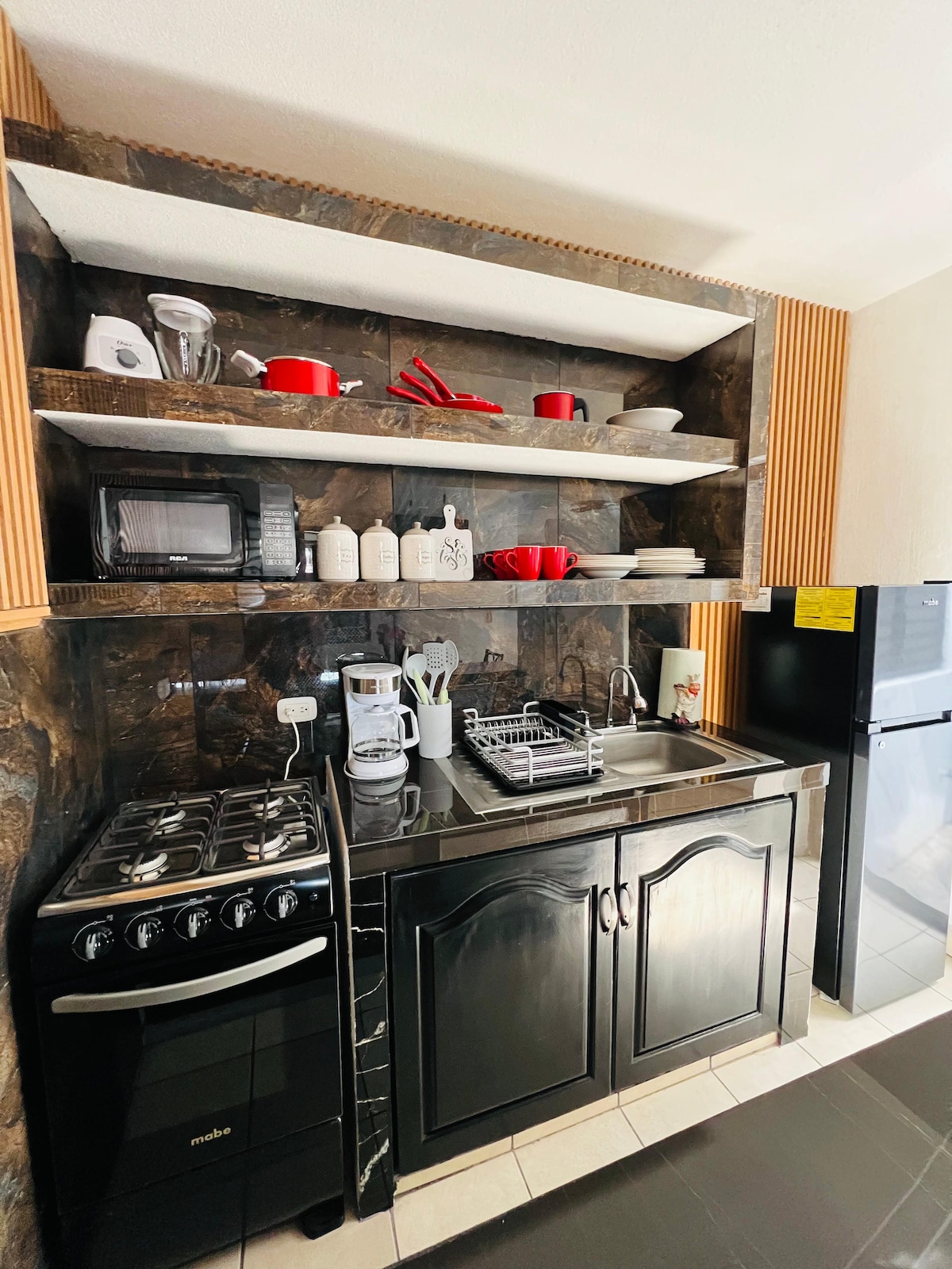 A compact kitchen area displays dark cabinetry and modern appliances. A gas stove, microwave, and refrigerator are visible, with white dishes and red accents arranged on open shelves above the countertop. The space is neatly organized for cooking and meal preparation.