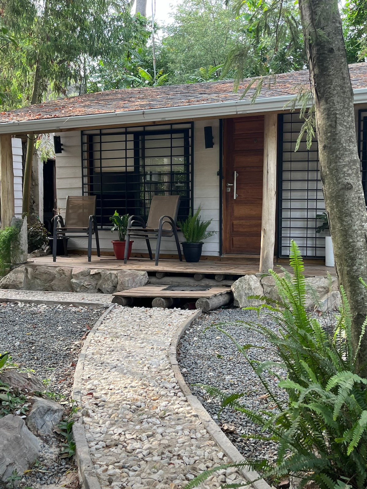 A welcoming entrance features wooden steps leading to a porch adorned with two comfortable chairs. Lush greenery surrounds the area, complemented by a flat stone pathway guiding visitors. Large windows offer a view into the cozy interior, while a wooden door adds rustic charm.