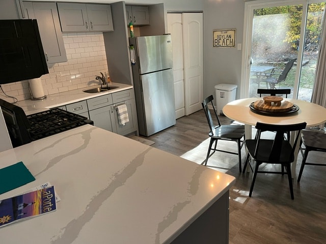 A modern kitchen space features gray cabinetry, stainless steel appliances, and a white countertop with a marble pattern. A small dining table with black chairs is positioned near a sliding glass door that opens to a backyard area with greenery.