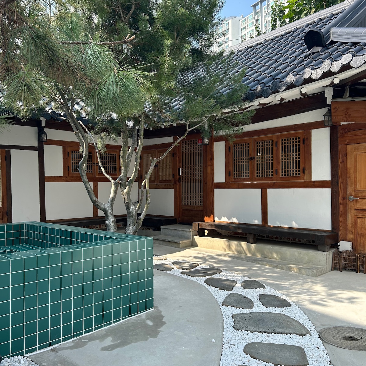 A traditional hanok courtyard is presented, featuring a lush green tree and a geometric tile water feature. The space includes stone pathways and wooden bench seating, creating a serene environment with harmonious architectural elements.