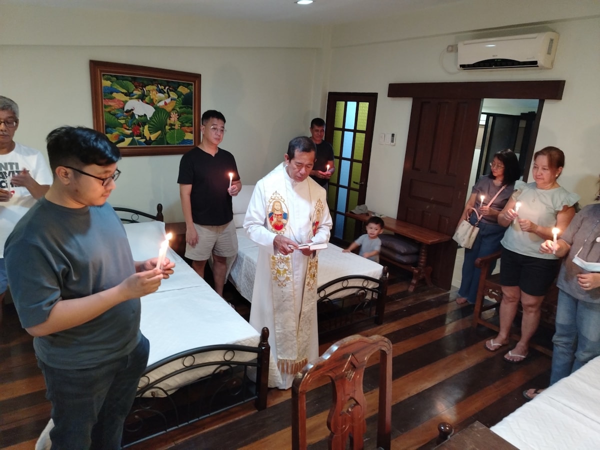 A bright room is filled with individuals holding candles. The center features a person in ceremonial attire. Two beds are positioned with white linens, and a wooden dining table is visible nearby. Natural light enters through windows, illuminating the gathering.
