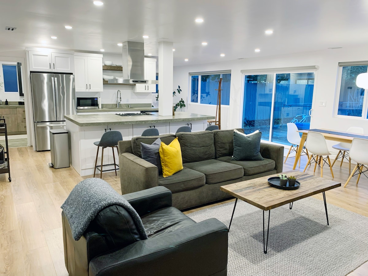 An open floor plan reveals a modern living area with a comfortable dark sofa and armchair. A coffee table centers the space, surrounded by a light area rug. The adjoining kitchen, featuring stainless steel appliances, is visible. Large windows provide natural light and views of the outdoor patio.