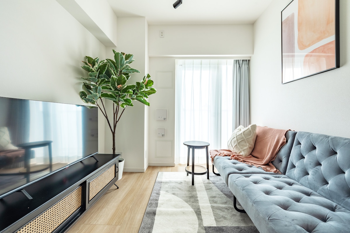 A modern living area features a sleek flat-screen TV positioned on a stylish media console. A grey fold-out sofa is complemented by a light-colored throw and cushion. A round table sits nearby, and a tall, leafy plant adds a touch of greenery next to the floor-to-ceiling window.