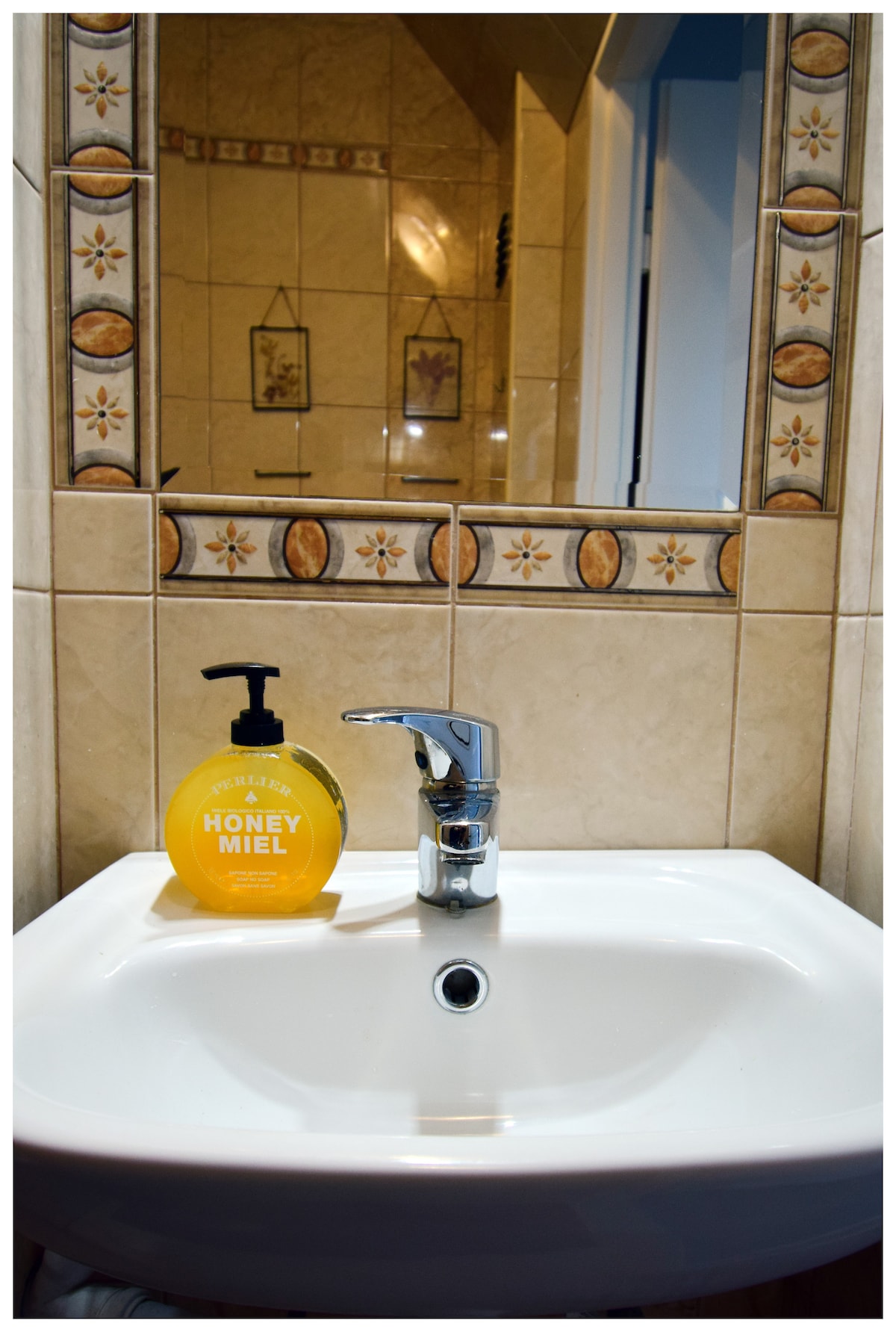 A clean bathroom sink is featured, accompanied by a pump bottle of honey-scented liquid soap. The sink is set against tiled walls with decorative floral accents, while a mirror reflects the surrounding area. Soft lighting enhances the serene atmosphere of the space.
