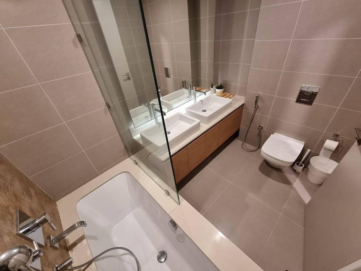 A modern bathroom is displayed, featuring dual sinks with sleek faucets and wooden cabinetry below. A glass shower enclosure is visible alongside a white bathtub. A toilet is positioned discreetly, and neutral tiles provide a clean and organized appearance.