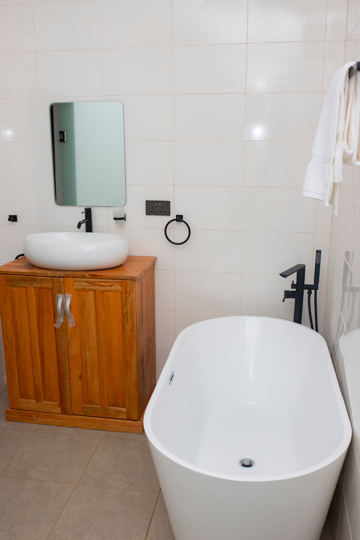 A modern bathroom features a freestanding white tub beside a wooden vanity topped with a round basin. A wall-mounted mirror reflects the natural light, while a black faucet and towel rack provide functional accents. Neutral tiles and a clean layout enhance the serene ambiance.