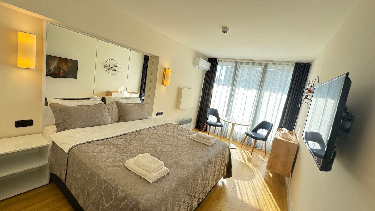 A spacious studio apartment features a king-size bed with soft linens, accompanied by large windows dressed in sheer curtains, allowing natural light. Two modern chairs and a small table are positioned near the window. A flat-screen TV is mounted on the wall opposite the bed.