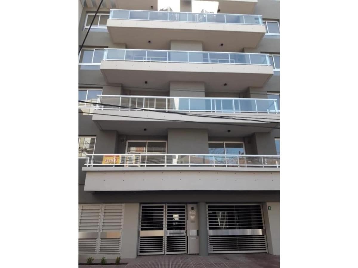 A modern building exterior is displayed with multiple floors and glass balcony railings. The entrance features a sleek design, and the façade is characterized by neutral colors and clean lines. Large windows allow for natural light, creating an inviting appearance.