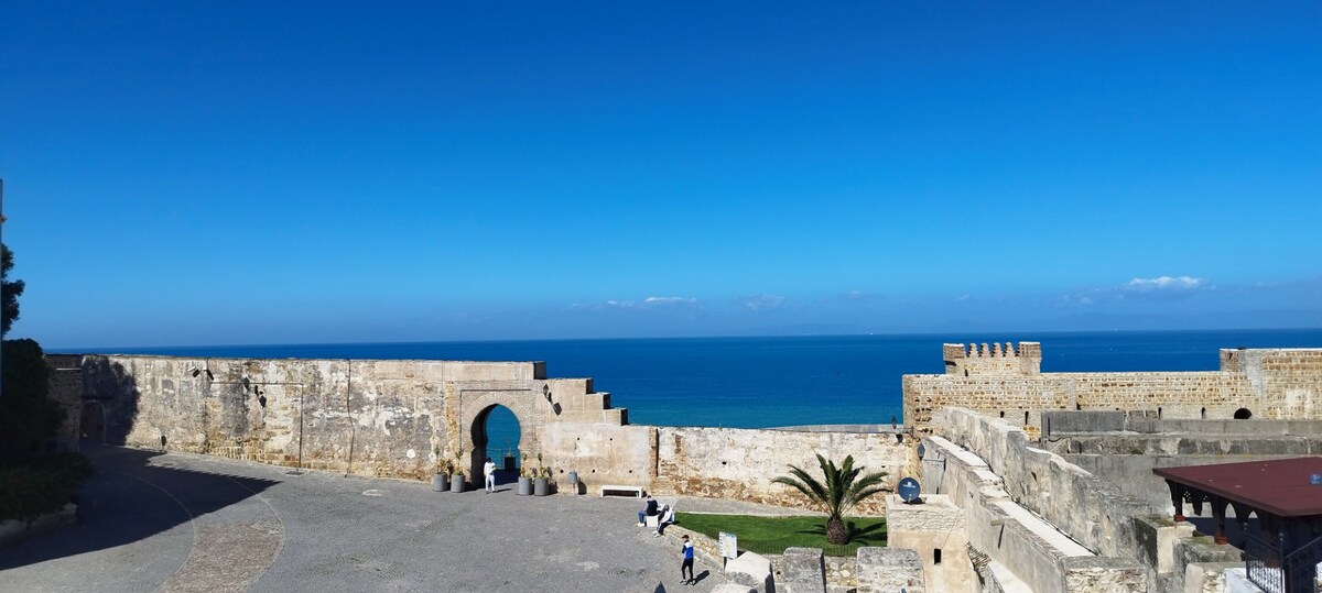 A coastal view features an ancient stone fortification overlooking the ocean. The clear blue sky contrasts with the weathered walls, and visitors are seen walking along the promenade. Palms are planted nearby, enhancing the serene outdoor atmosphere.