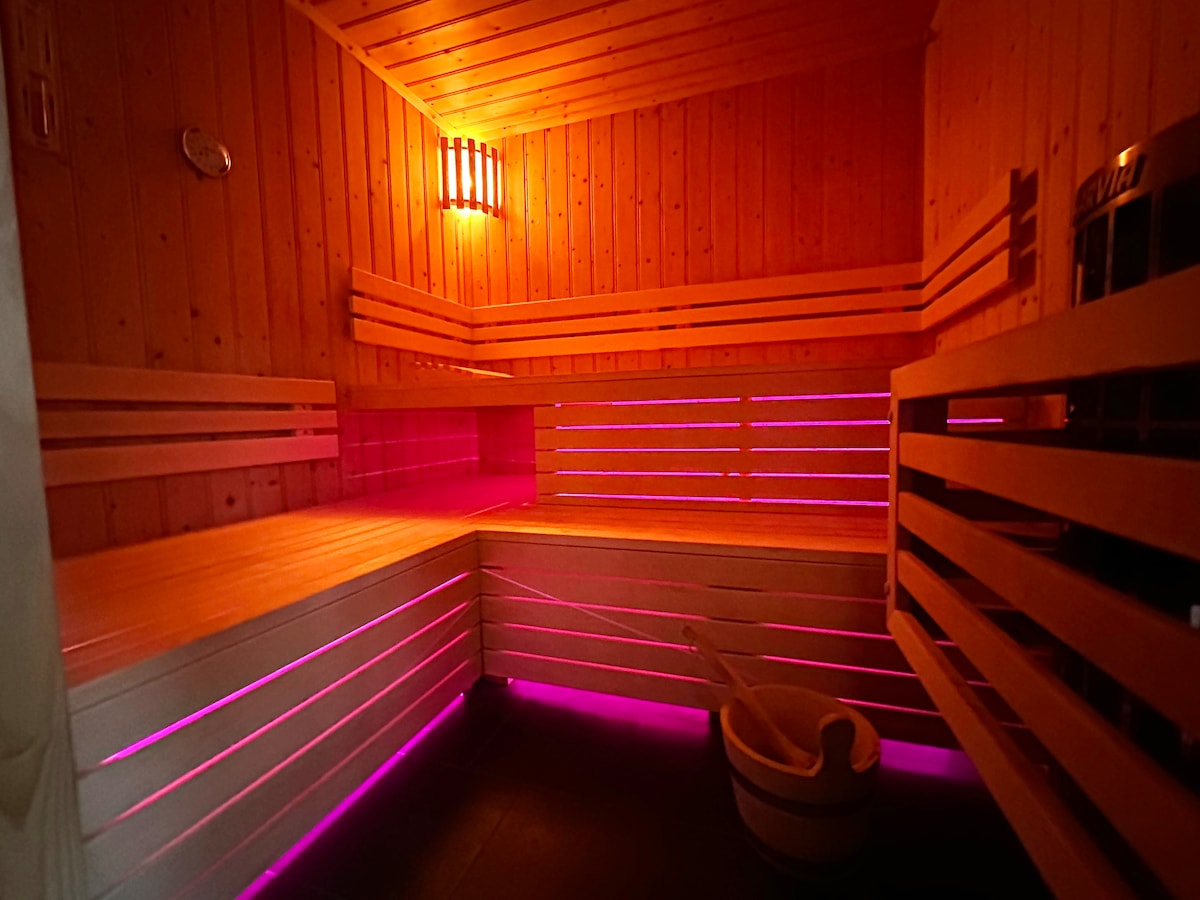 A private sauna room features wooden paneling and multiple benches. Soft lighting in purple hues highlights the ambiance, and a traditional wooden bucket is positioned at the side. The inviting space offers a serene environment for relaxation.