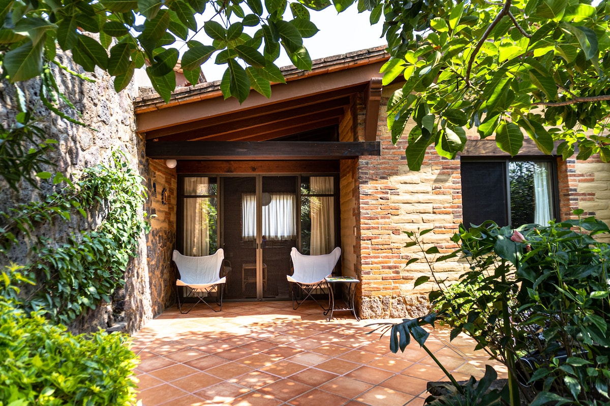 A private patio is framed by lush greenery, featuring two comfortable chairs positioned invitingly. The entrance to the cabin, with brick walls and wooden beams, is partially visible, complemented by the warm tones of the tiled flooring.