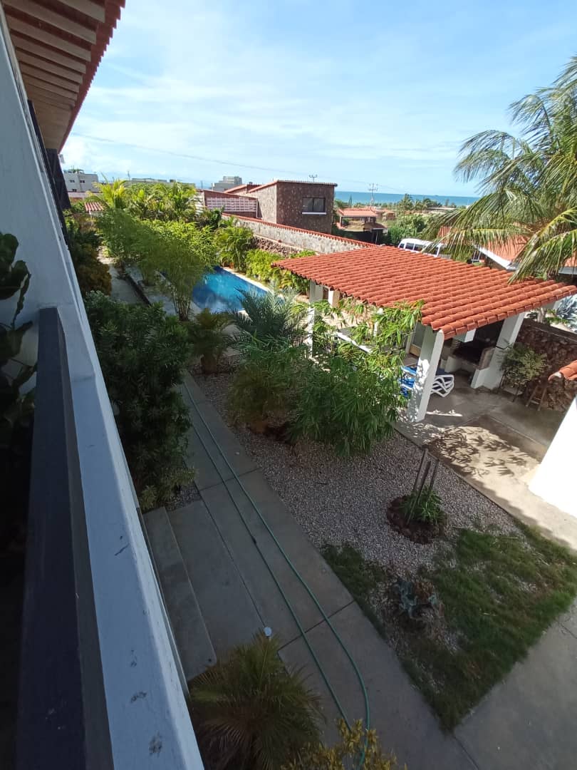 A view from the second floor showcases a garden area with a pathway lined by greenery and palm trees. The pool area is visible in the distance, accompanied by terracotta-roofed structures. A serene atmosphere is created by abundant natural light and open spaces.
