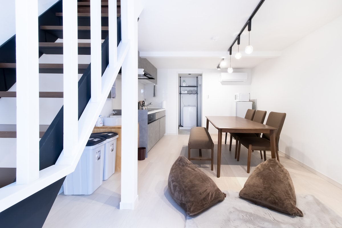 The living and dining area features a spacious table surrounded by four chairs, with two plush floor cushions nearby. A well-equipped kitchen is visible in the background, alongside a laundry area. Natural light enhances the neutral-toned interior, complemented by wooden accents.