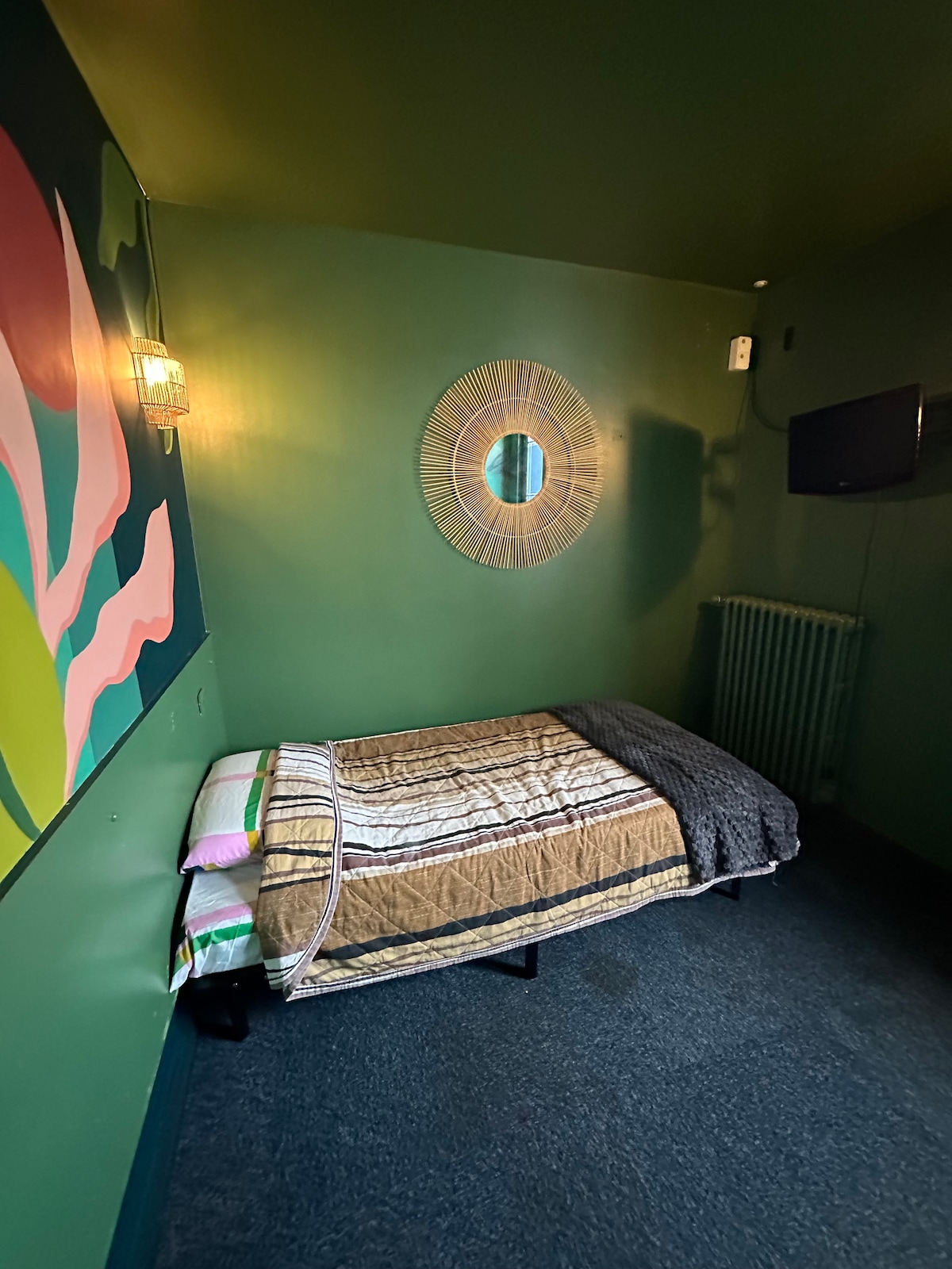 A small room features a single bed dressed with a patterned blanket and a soft throw. The walls are painted a deep green, complemented by a large wall art piece. A round decorative mirror is positioned above the bed, and a radiator is visible nearby.