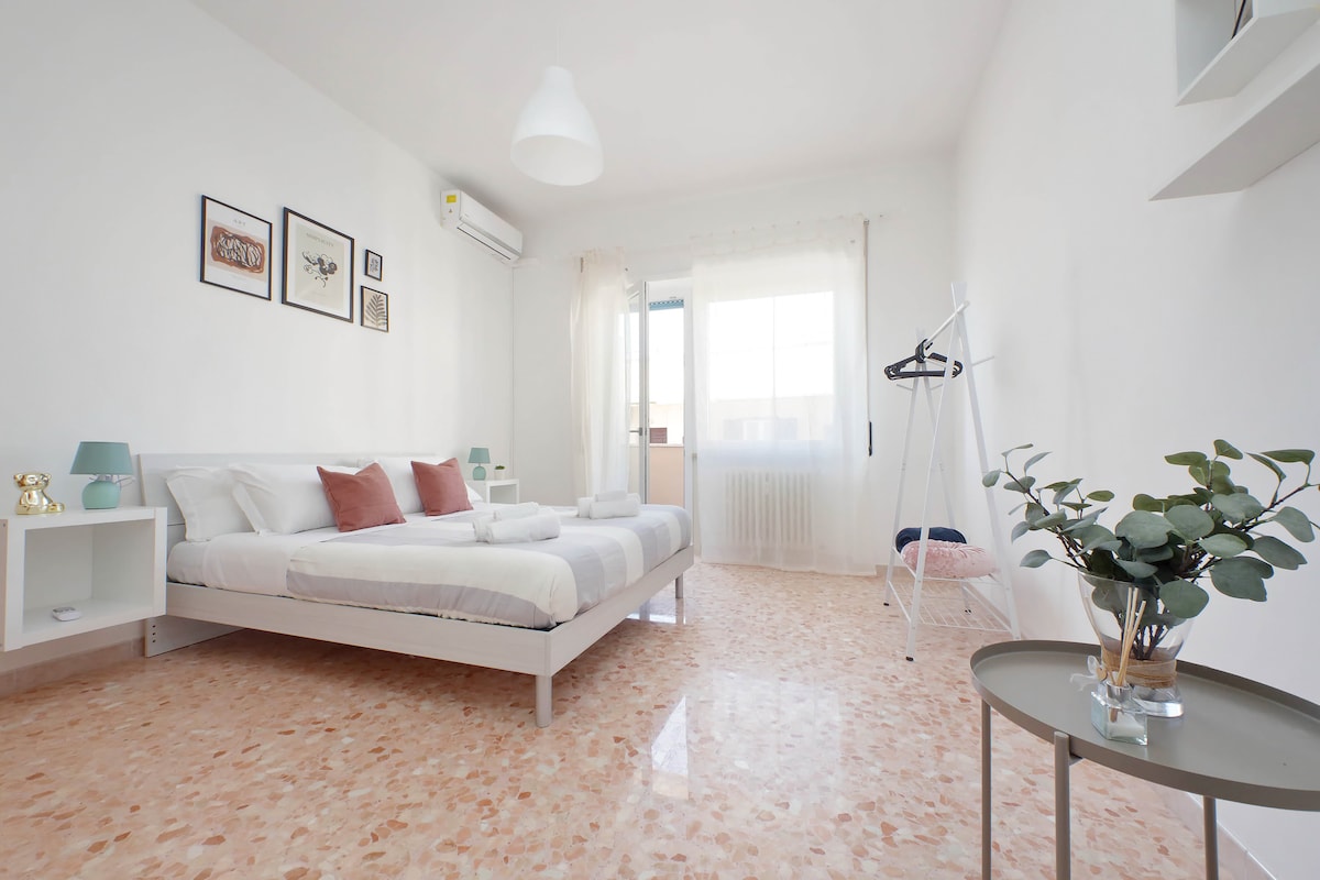 The sleeping area features a spacious bed with multiple pillows and a light-colored blanket. A minimalist design is accentuated by framed artwork on the walls. Natural light filters in through the window, illuminating a light-colored floor. A small table holds a potted plant, adding a touch of greenery.