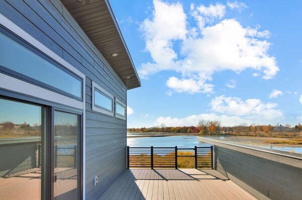 The image displays a modern exterior patio with wooden decking, bordered by a sleek railing. Large glass doors provide a view of the serene water and surrounding landscape, with patches of blue sky and soft clouds above.