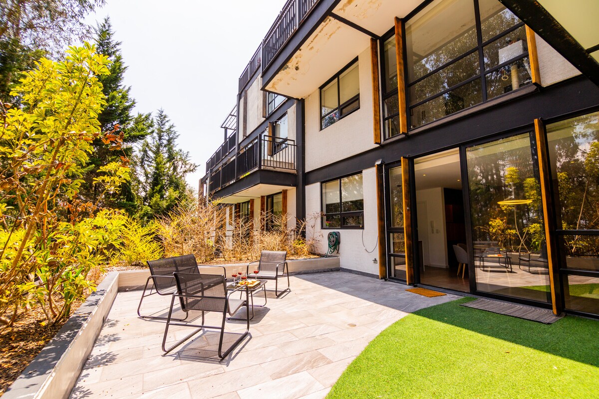 A spacious outdoor patio area is surrounded by greenery, featuring a well-maintained lawn and stone flooring. Several lounge chairs are arranged around a small table, providing a comfortable space to relax and enjoy the natural surroundings.
