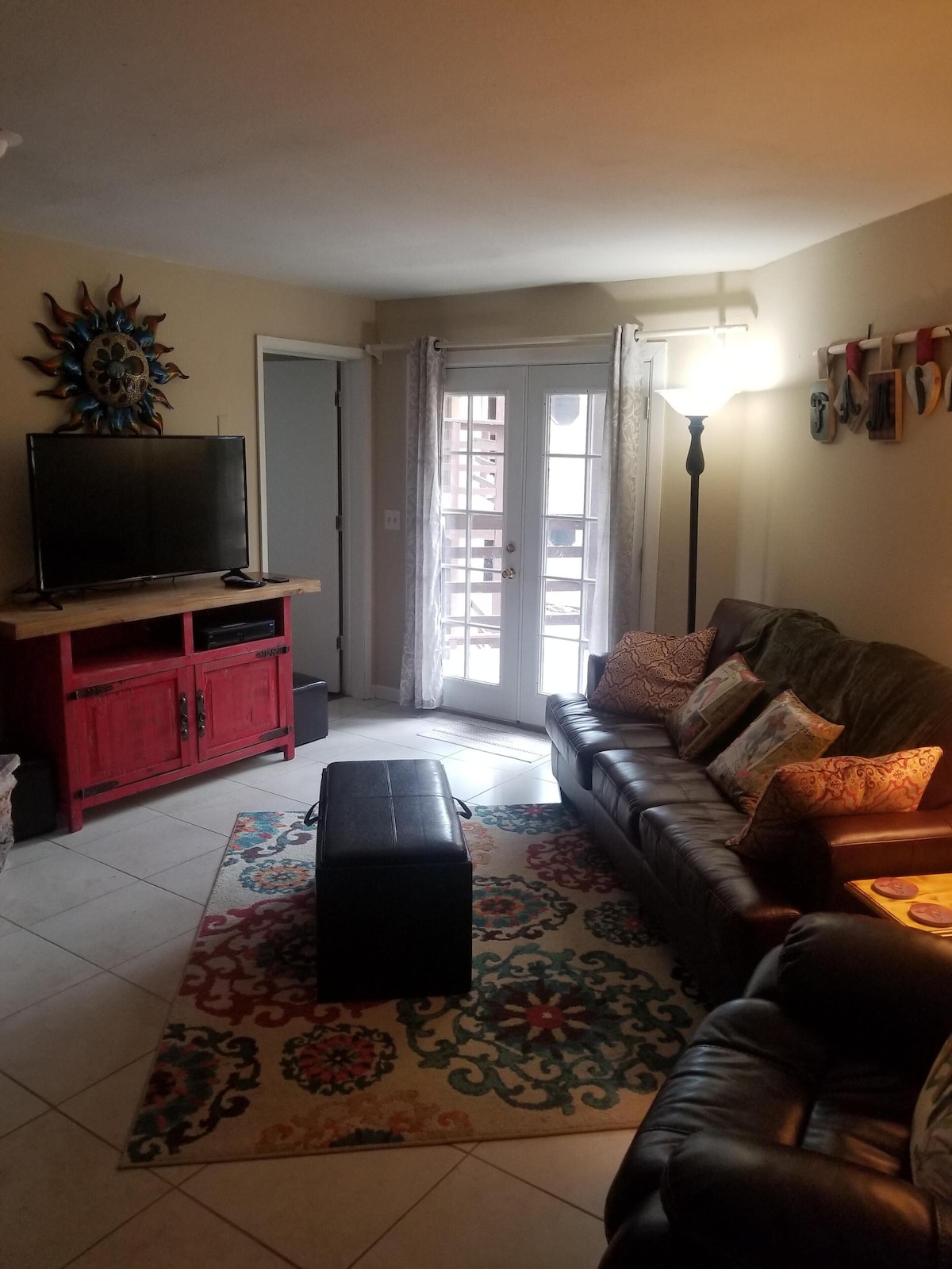 A welcoming living area features a leather sofa adorned with decorative pillows, positioned near a wooden entertainment unit with a television. A colorful area rug covers the tiled floor, while natural light enters through glass doors leading to a small outdoor space.