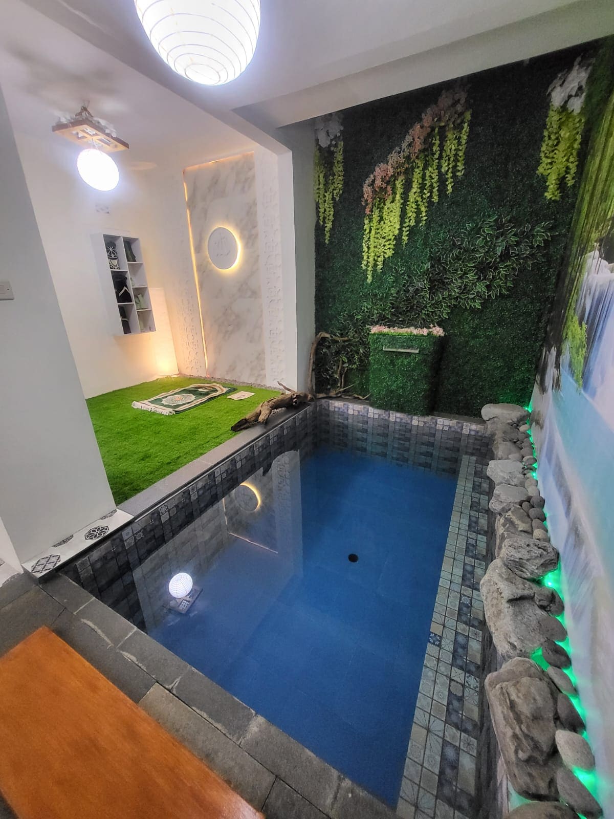 A tranquil indoor pool is framed by natural stone and vibrant green wall decor. Soft lighting highlights the clear blue water. A wooden bench is positioned nearby, and an accent wall featuring a waterfall scene enhances the serene ambiance.