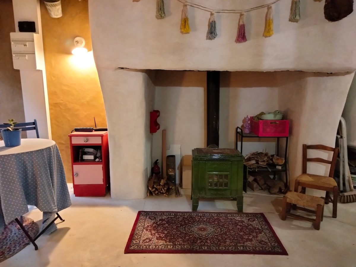 A compact living space features a cozy fireplace inset into a curved wall. A green wood-burning stove sits on a beige floor, surrounded by logs and decorative items. A small round table with a blue tablecloth and a plant is visible, alongside a stack of wooden chairs.