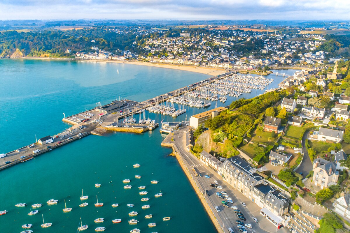 An aerial view captures a serene coastal landscape with a marina filled with boats. The beach is lined with sand and dotted with people. Lush green hills rise in the background, and residential buildings provide a scenic contrast to the sparkling blue water.