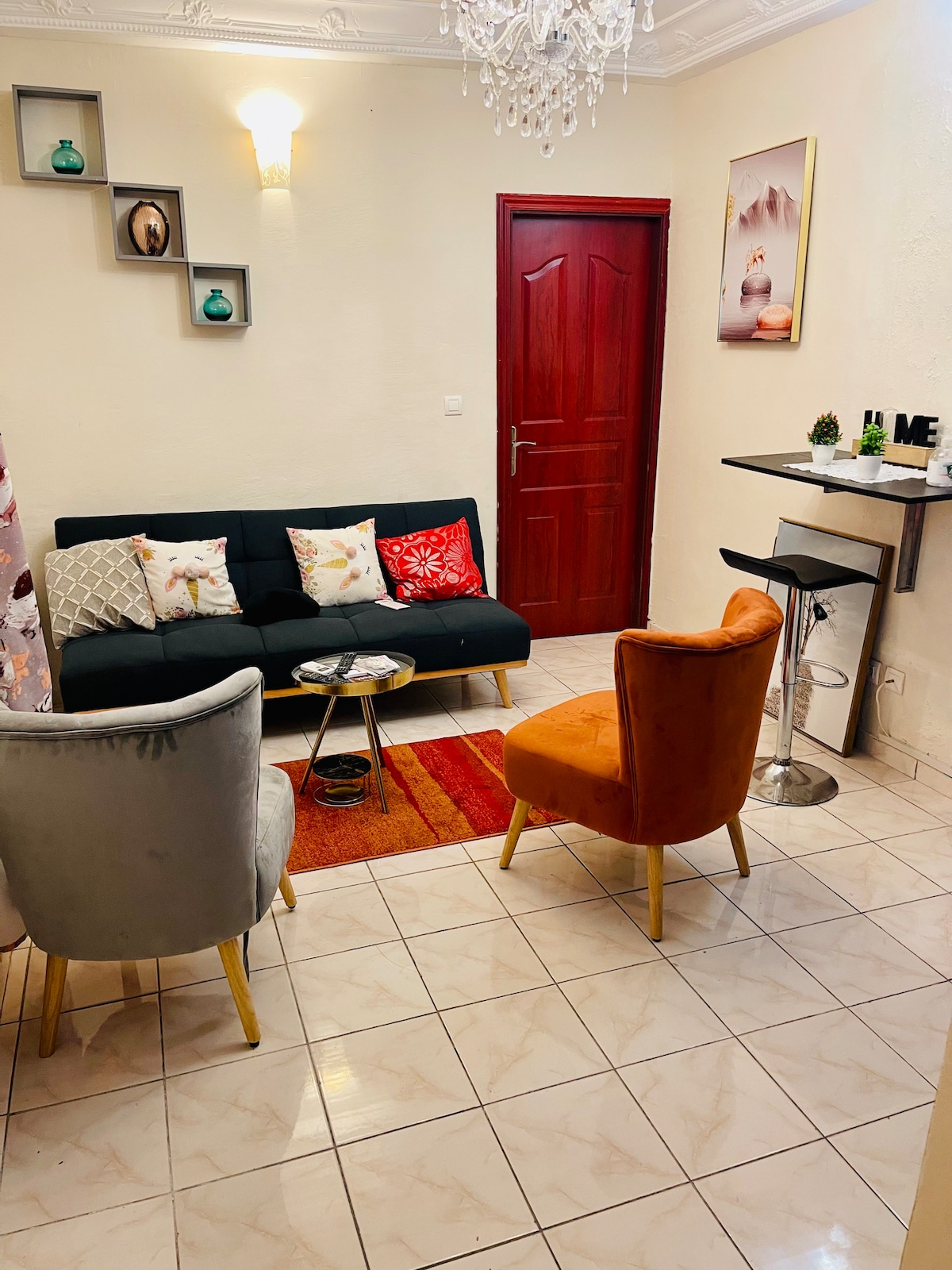 A cozy living area is furnished with a black sofa and two accent chairs in gray and orange. Decorative cushions add color, while a small table holds a few items. The space features a small bar table against the wall, enhancing the inviting atmosphere.