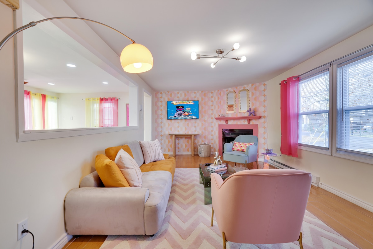 A cozy living room is featured, showcasing a plush sofa with colorful cushions, complemented by a pink armchair. A wall-mounted TV is positioned above a decorative fireplace, while large windows enhance natural light, accented by sheer curtains in vibrant colors.