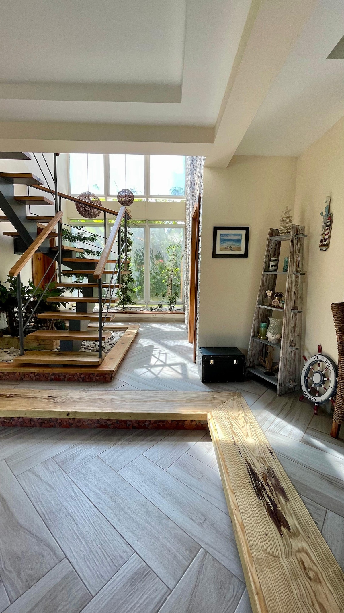 A bright entrance area is featured, showcasing an open staircase made of wood and metal. Natural light pours in through large windows, illuminating the space. Decorative plants and art pieces add a touch of warmth, while wooden benches offer seating along the floor.
