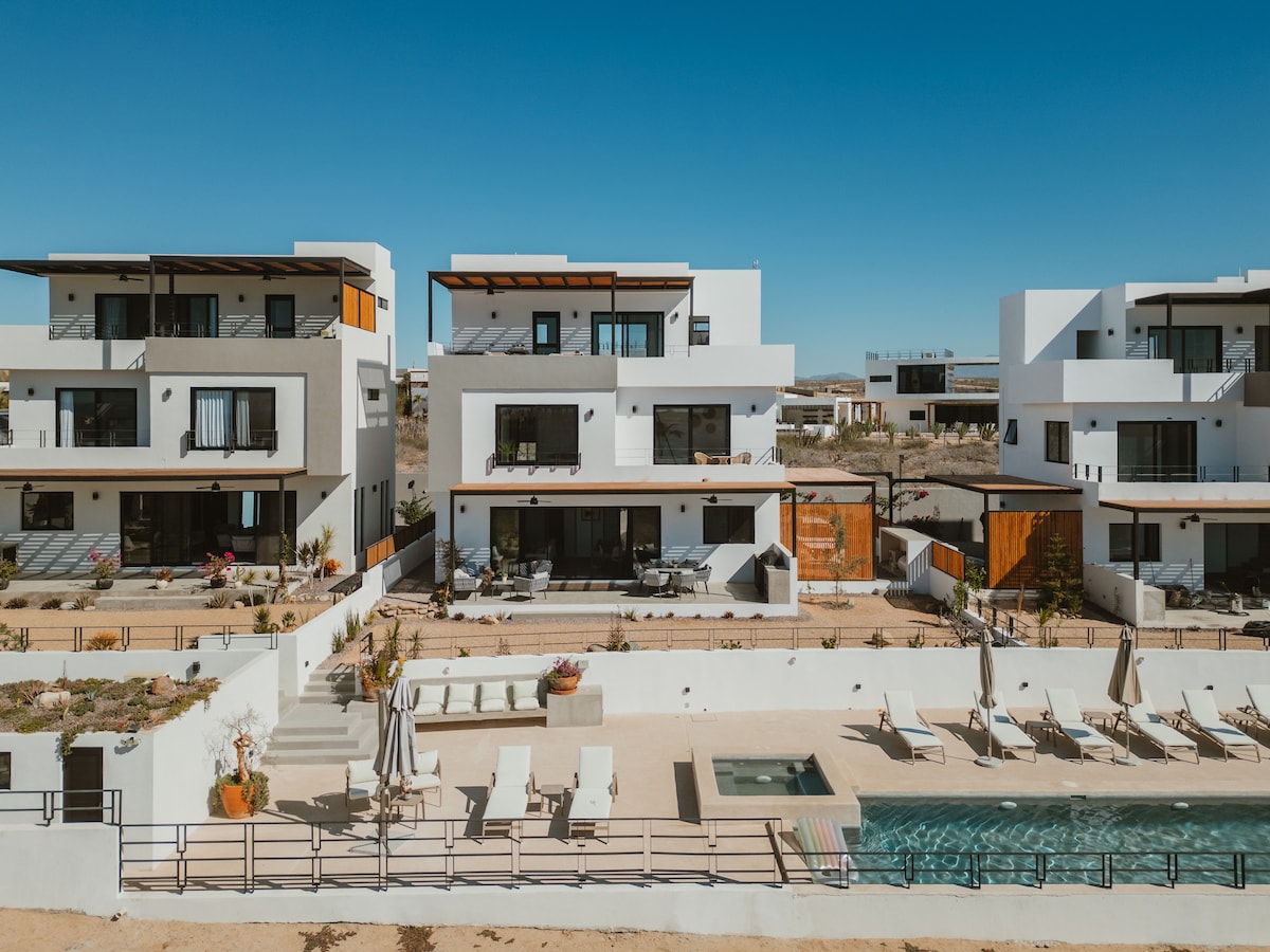 A row of modern homes features white exteriors and large windows. A spacious shared pool is visible in the foreground, surrounded by sun loungers. The outdoor area is designed for relaxation and enjoyment, with decorative plants scattered throughout the landscaped grounds.