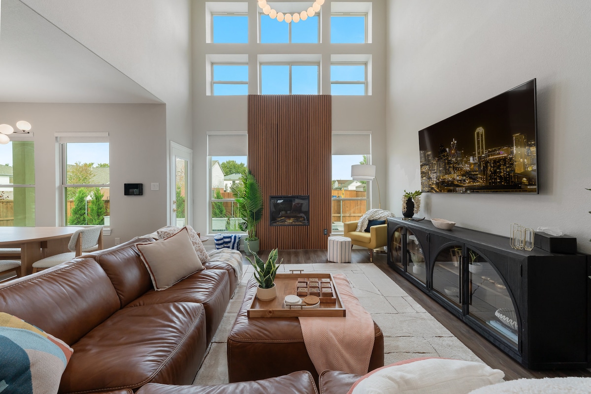 A modern living area features large windows that fill the space with natural light. A comfortable sectional sofa is positioned in front of a stylish media console. A fireplace is centrally located, complemented by a wall art piece, creating a cozy focal point in the room.
