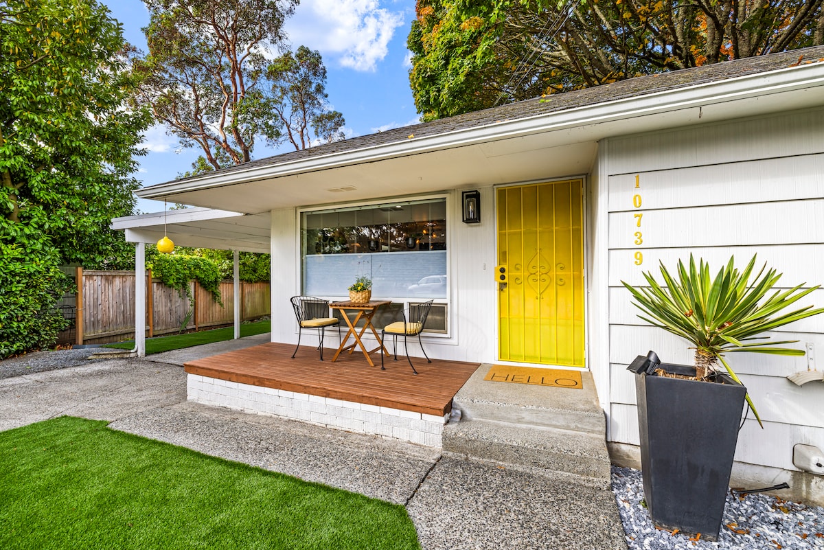 A welcoming entrance features a bright yellow door and a small, covered front deck with a table and two chairs. Lush green grass borders the area, complemented by a potted plant. A pathway leads to the carport, framed by tall trees for added privacy.