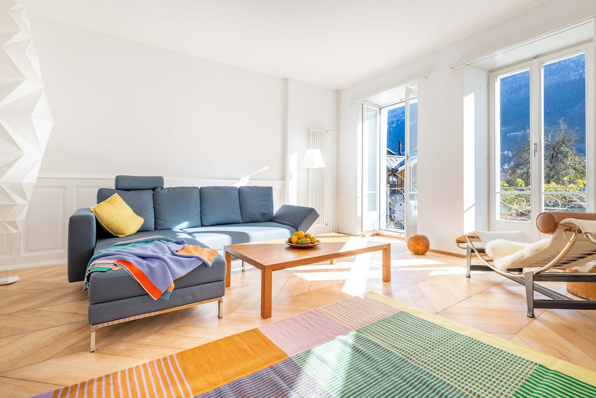 A bright living area features a modern blue sofa adorned with a colorful throw. A wooden coffee table is positioned on a vibrant area rug, with sunlight illuminating the space through large windows that offer mountain views.