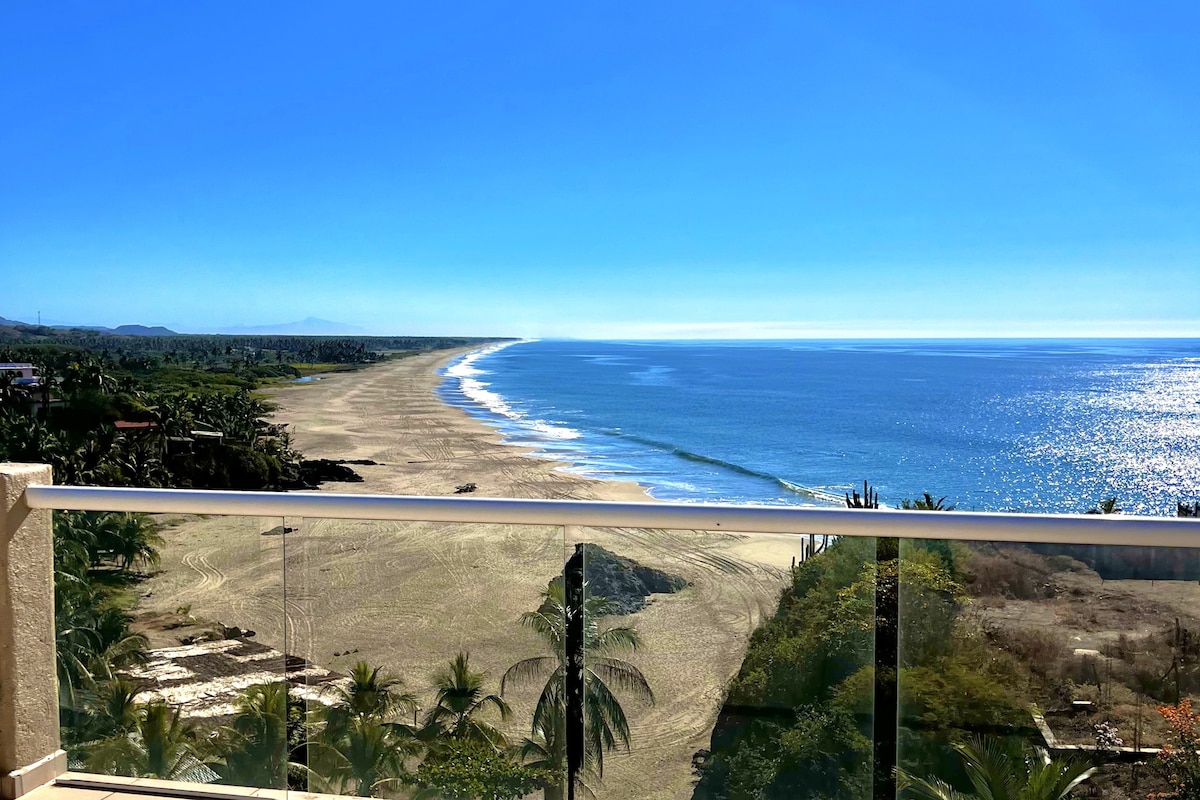 A panoramic view of the coastline is captured, showcasing sandy beaches meeting the ocean under a clear blue sky. Lush greenery lines the shore, while gentle waves reflect the sunlight, creating a serene landscape that invites relaxation.