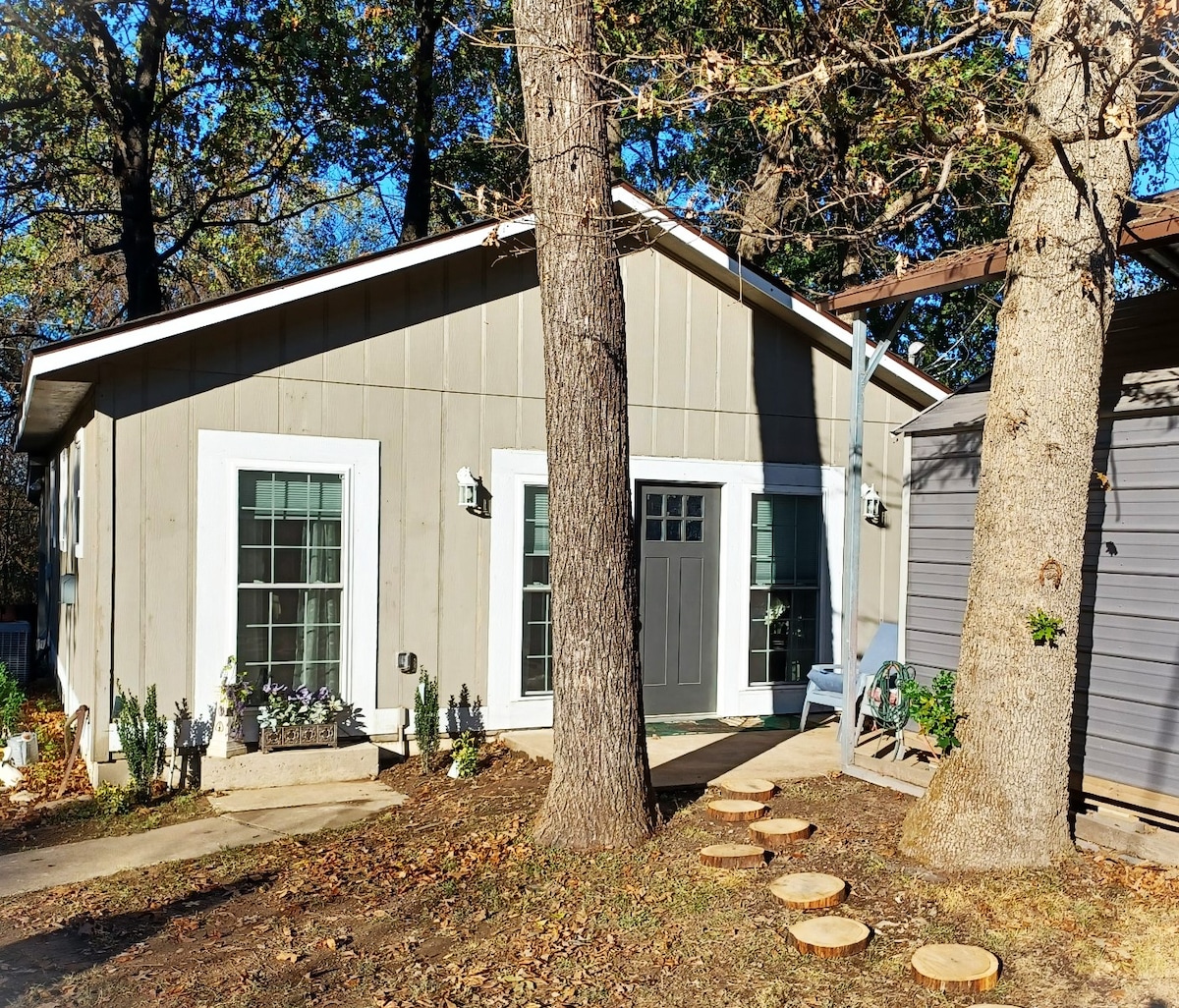 A newly constructed guest house is shown, featuring a light gray exterior and large front windows. A welcoming front door is flanked by two outdoor light fixtures. Stone steps lead to the entrance, surrounded by trees and landscaped with a few potted plants.