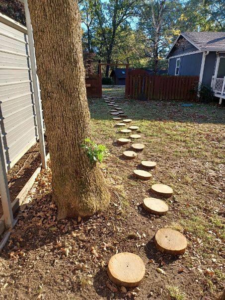 A thoughtfully laid stepping stone path winds through a grassy yard, bordered by trees. The circular stones vary in size and are positioned evenly, leading towards a wooden fence. The surrounding area is decorated with fallen leaves, creating a natural and serene setting.