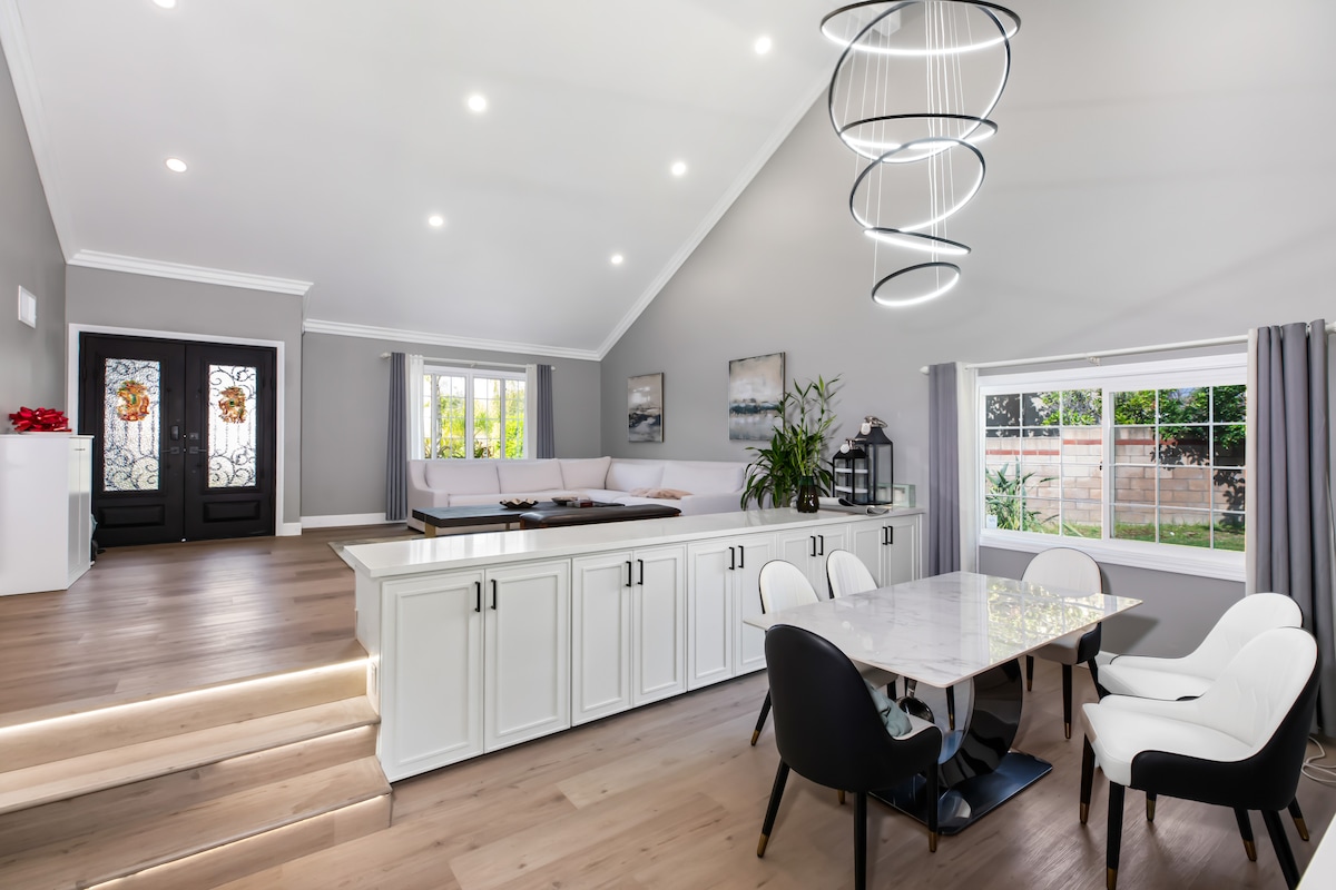 An open-concept living area featuring a high ceiling. A modern dining table with black and white chairs is positioned near a sleek kitchen island, illuminated by a stylish light fixture. Natural light filters through large windows, enhancing the airy space.