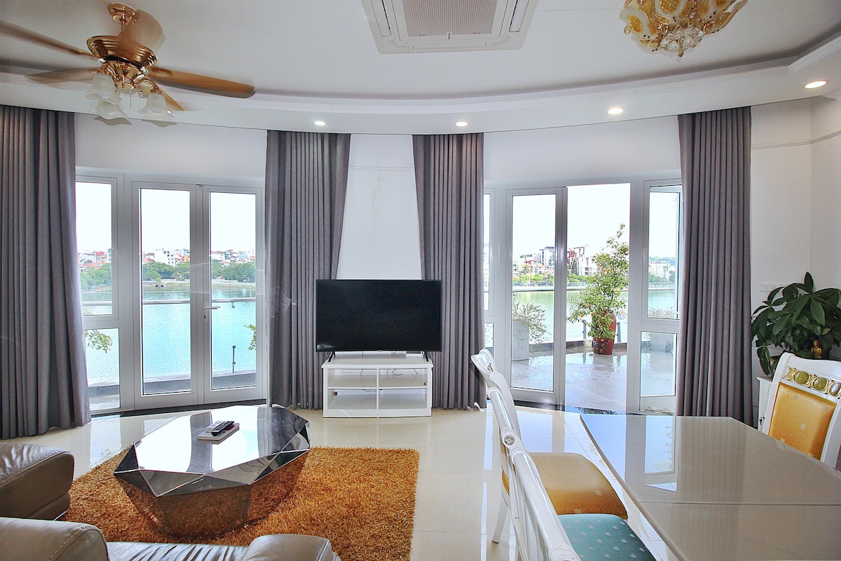 A spacious living area features large windows that offer views of the lake. A flat-screen television is mounted on a white cabinet, while a crystal chandelier adds elegance. Light grey curtains frame the windows, and a modern coffee table sits on a soft area rug.