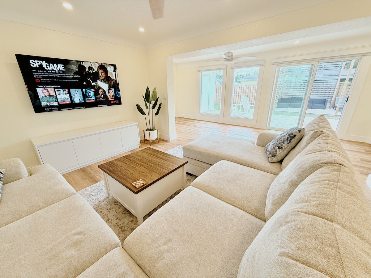A light and airy living area features a spacious sectional sofa in soft beige, complemented by a wooden coffee table. A large flat-screen television is mounted on the wall, displaying movie options. Sunlight filters through sliding glass doors that lead to an outdoor space.