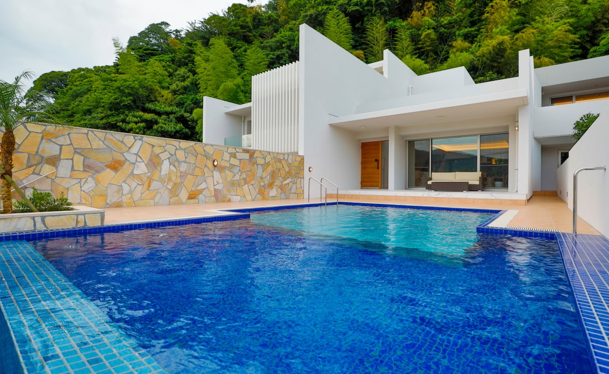 An outdoor private pool is surrounded by a stone wall and lush greenery. The crystal-clear water reflects the nearby architecture, featuring a spacious patio area with modern style and large glass windows. Steps lead gently from the pool to the living area.