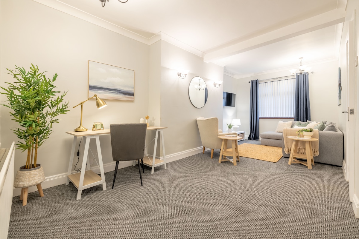 A spacious living area features a soft grey sofa and neutral decor, complemented by a round mirror and modern lighting. A work desk with a chair and a small plant add functionality. Natural light filters through a large window, illuminating the welcoming space.