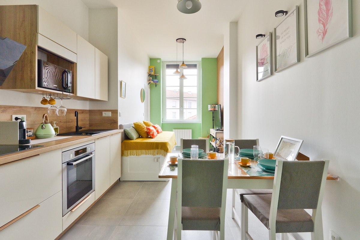 The open kitchen features modern cabinetry and appliances, including a refrigerator and oven. A comfortable seating area with a daybed is positioned near a large window. A dining table is set for meals, surrounded by inviting chairs and decorated walls with framed art.