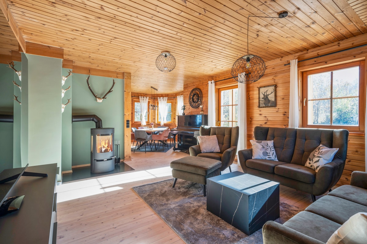 An inviting living area features wooden walls and a ceiling, with large windows allowing natural light. A cozy seating arrangement includes two dark sofas facing a stylish coffee table. A wood stove adds warmth, while a dining area is visible in the background.