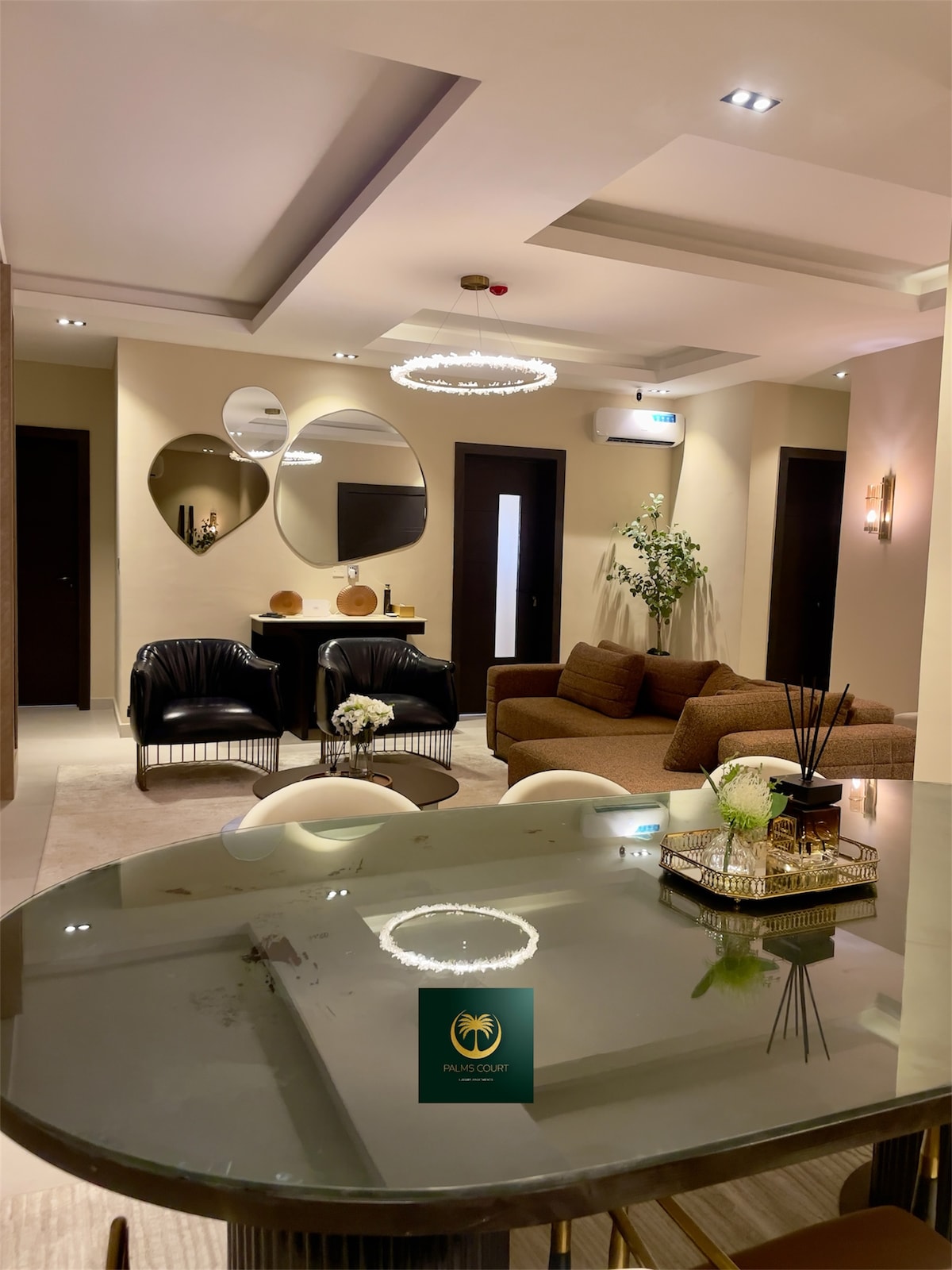 The living area is characterized by modern design elements, featuring a comfortable seating arrangement with a brown sofa and two black chairs. A glass dining table is positioned nearby, illuminated by a chandelier. Mirrors on the wall enhance the spacious ambiance.