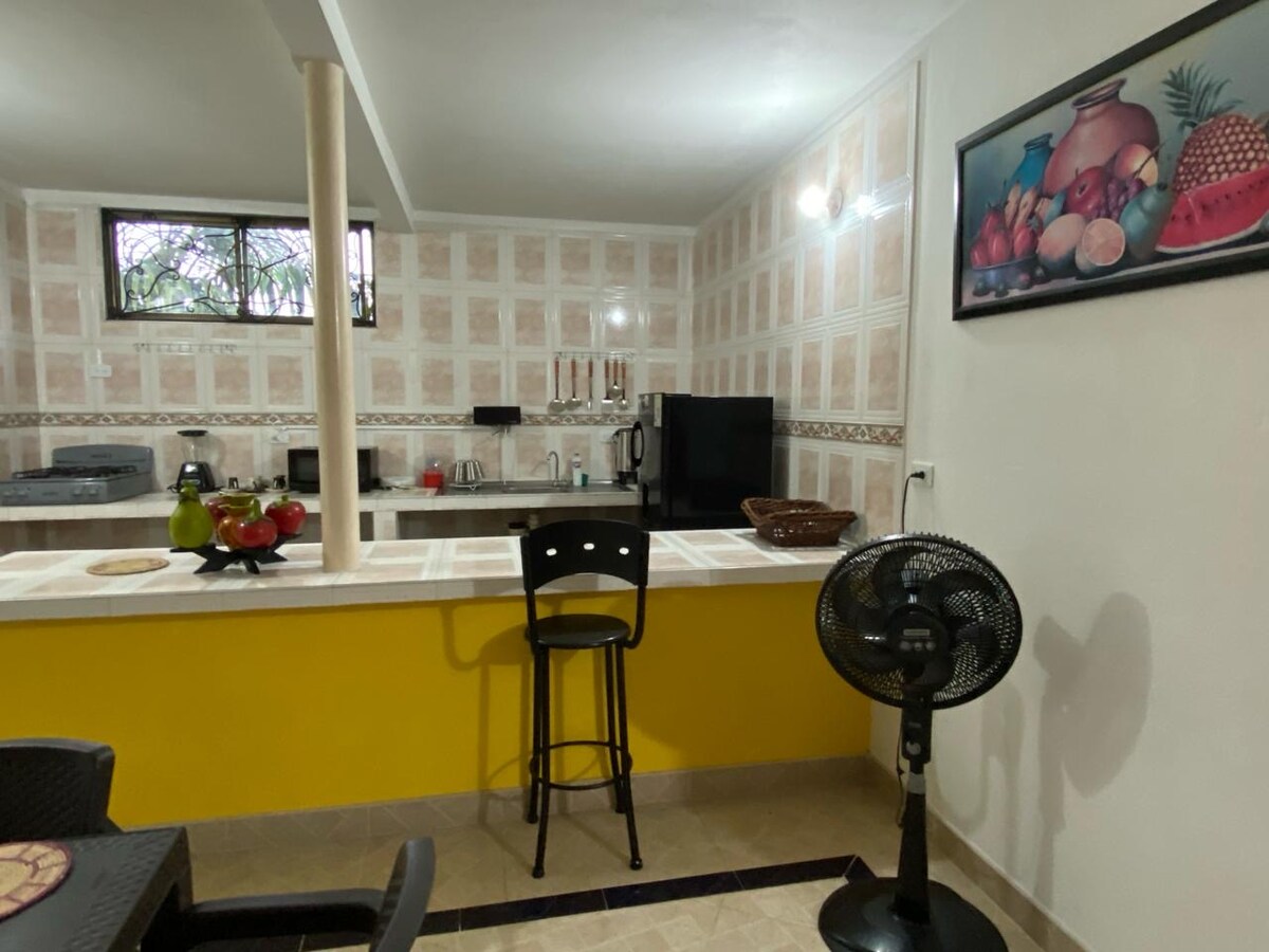A bright kitchen area is highlighted by light-colored tiled walls and a yellow accent bar. A kitchen counter with two stools provides seating, while a variety of appliances and a television are visible in the background. Natural light enters through the window, creating a welcoming environment.