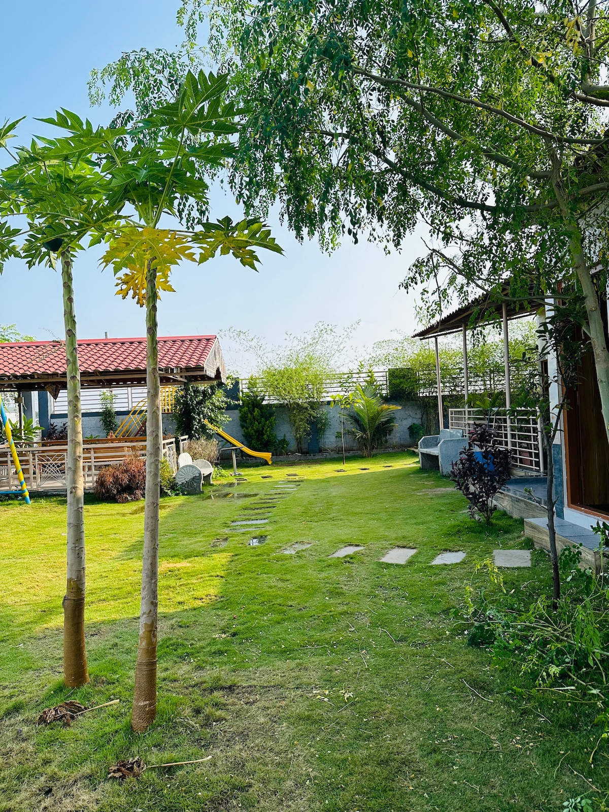 A lush green garden is framed by young trees, featuring a sunlit path of stepping stones leading towards a shaded seating area. A traditional gazebo is visible, accompanied by a hammock and manicured foliage, creating an inviting outdoor space for relaxation.