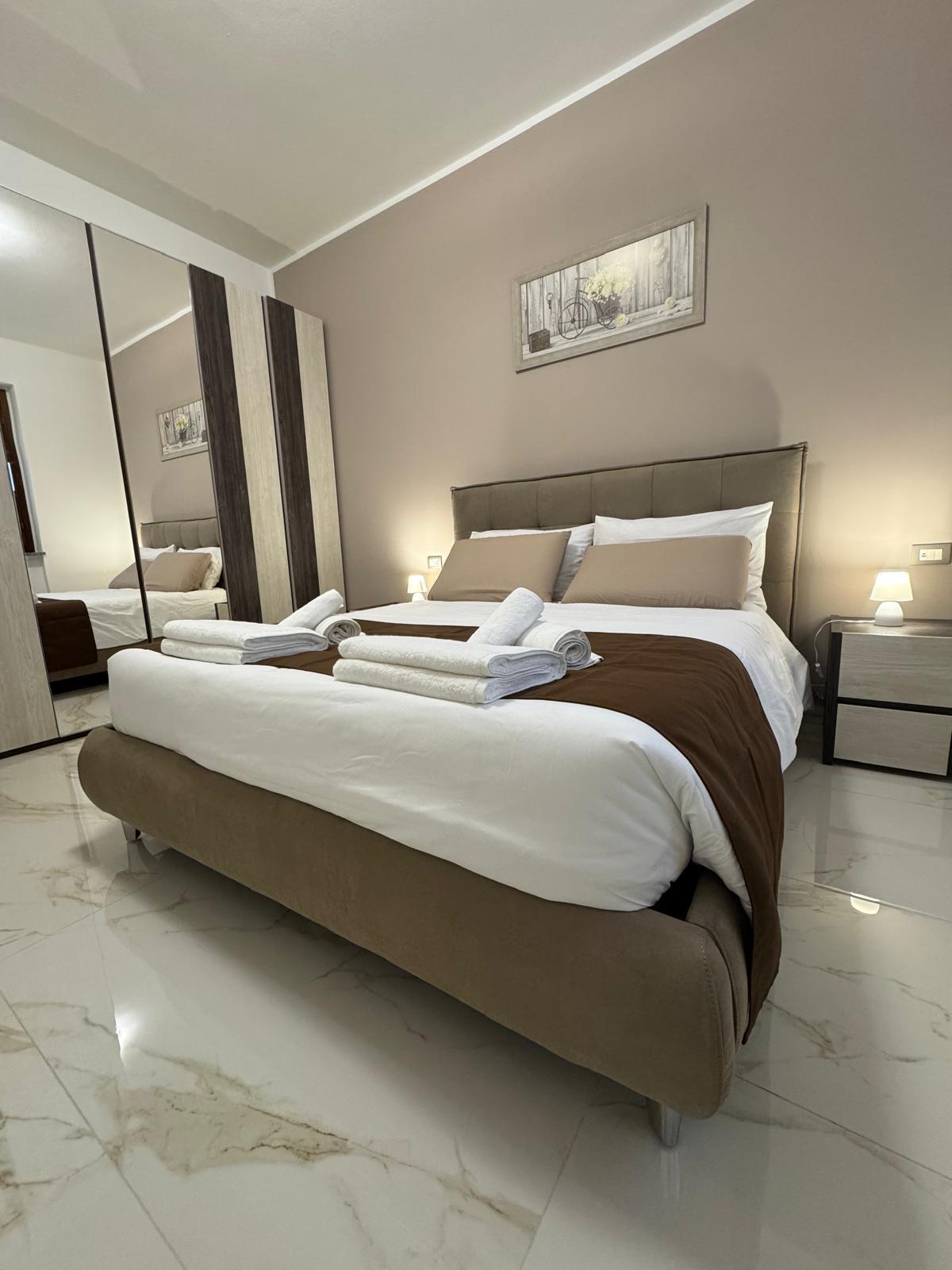 A modern bedroom features a spacious bed with a neutral-colored comforter and decorative pillows. Two neatly arranged towels are placed on the bed. A mirrored closet reflects the soft lighting provided by bedside lamps, while a framed artwork adds a subtle touch to the decor.