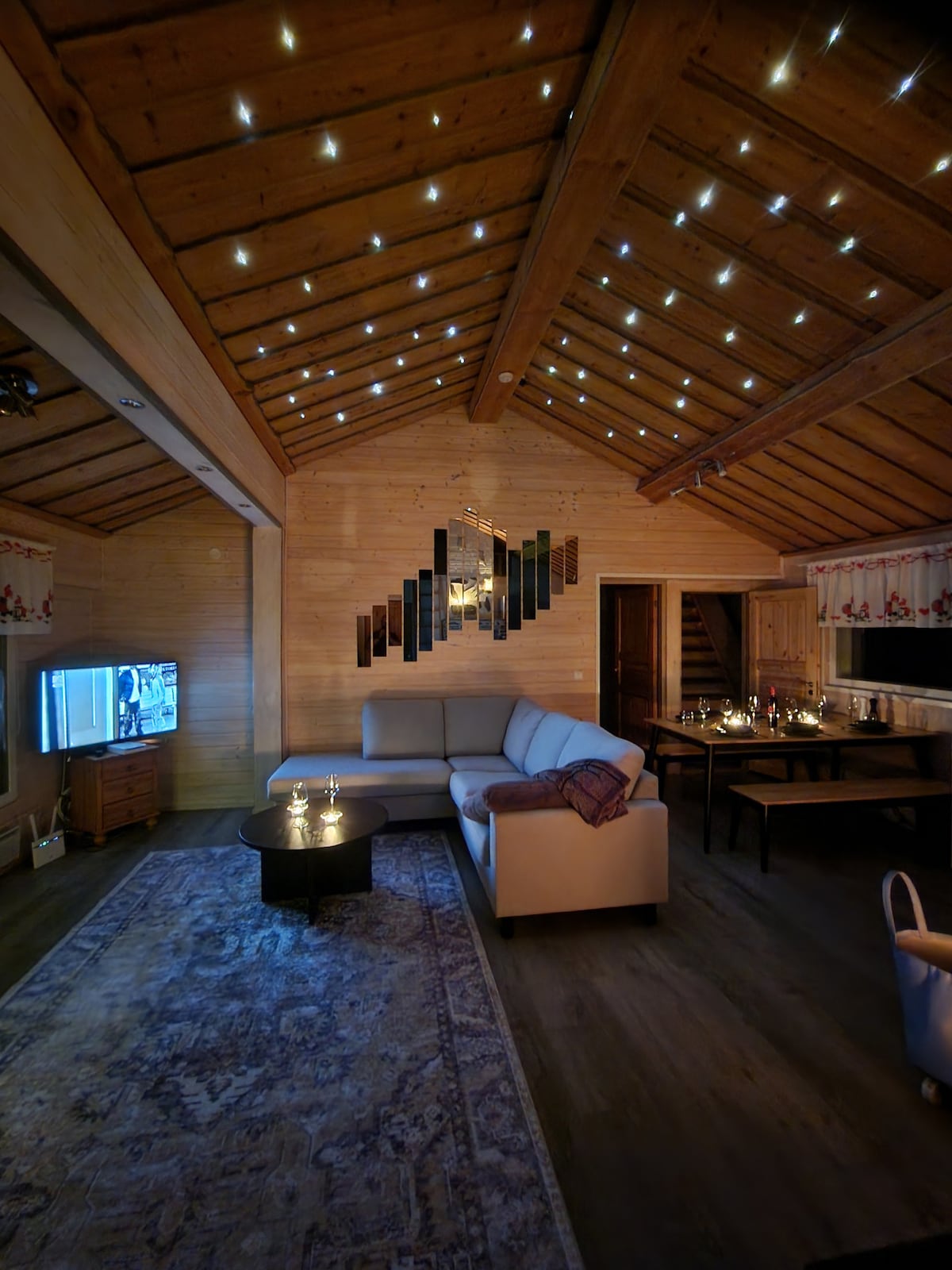 The living room features a high wooden ceiling adorned with star-like lights, creating a serene ambiance. A light-colored sofa is positioned around a circular coffee table, alongside a cozy dining area. The space showcases contemporary décor with a blend of wood and subtle patterns.
