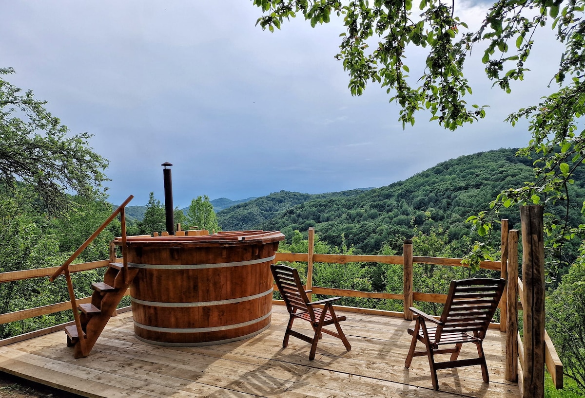 A wooden hot tub is surrounded by a spacious deck, complemented by two slatted lounge chairs. A scenic view of rolling green hills is visible in the background, framed by trees, providing a serene outdoor atmosphere.